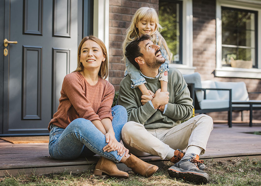 A mom and dad sitting on the floor of their front porch, a young daughter standing behind them, hugging her dad from behind. Do I Need Life Insurance?
