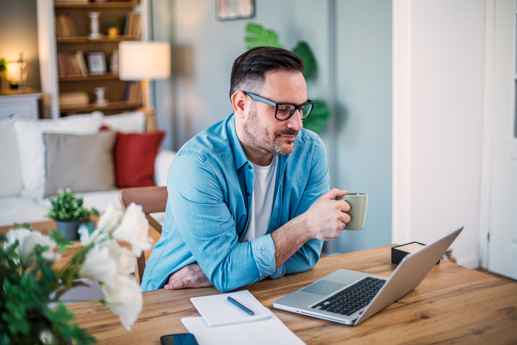 Handsome man sitting in home office, relaxing, positive, looking at laptop, drinking coffee, after work done.